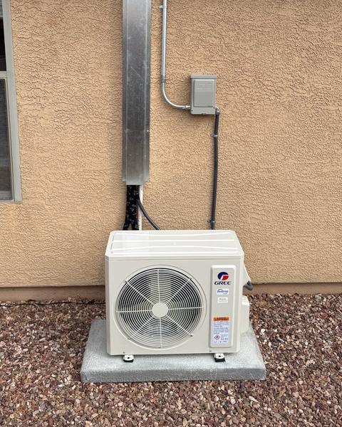 Gree outdoor mini-split on a pad beside a stucco wall, with a tall metal line cover and electrical disconnect